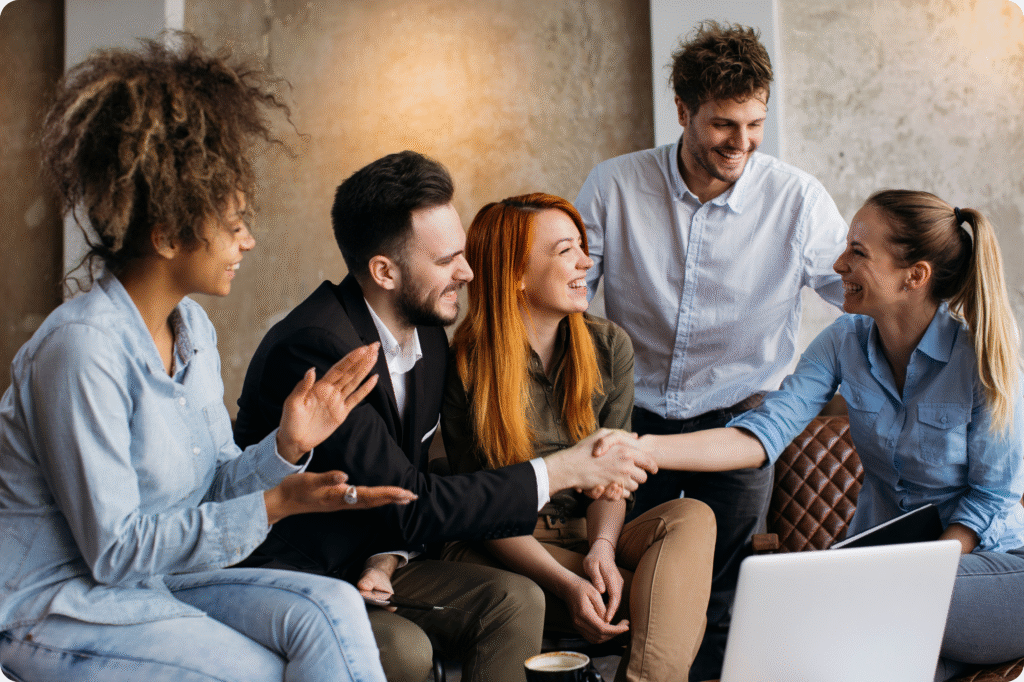 A diverse group of five professionals sitting together in a casual office setting. Two people are shaking hands and smiling, while the others engage in friendly conversation, suggesting collaboration and successful teamwork.